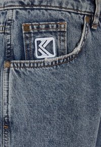 Blå denimjeans med en sliten textur, med en vit broderad logotyp på bakfickan och kopparfärgad hårdvara.