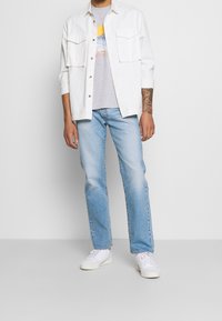 Camisa blanca de algodón sobre una camiseta gris con estampado, combinada con vaqueros de mezclilla azul claro y zapatillas blancas, posando sobre un fondo gris.