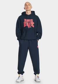 Granatowa bluza z kapturem z czerwonym nadrukiem "Prosto Athletic", z kieszenią kangurką, zestawiona z dopasowanymi spodniami dresowymi z akcentami logo. Białe buty sportowe.