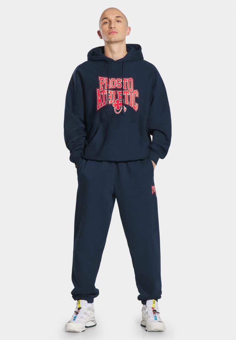 Granatowa bluza z kapturem z czerwonym nadrukiem "Prosto Athletic", z kieszenią kangurką, zestawiona z dopasowanymi spodniami dresowymi z akcentami logo. Białe buty sportowe.