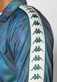 Camisa de manga larga de tonos azules y verdes con una franja blanca que presenta diseños de logotipos verdes repetidos a lo largo de la costura del hombro.
