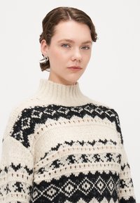 Cremfarbener Strickpullover mit schwarzen geometrischen Mustern, hohem Hals und strukturiertem Design. Das Model trägt einen silbernen Ohrring mit abstrakter Form.