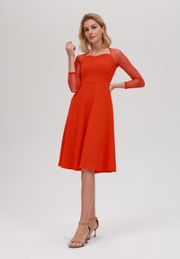 Robe rouge avec un corsage ajusté et une jupe évasée, dotée de manches longues transparentes à pois et d'un col en vagues. Tissu lisse.