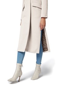 Long manteau beige avec poches avant, arborant une doublure en imprimé léopard, porté sur un jean bleu clair et des bottines beige clair.