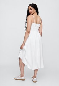 Vit, flödande klänning med en rynkad överdel, tunna axelband och en asymmetrisk fåll. Matchas med ljusa sneakers med gummisula.