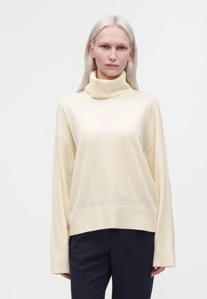 Femme à la peau pâle et aux cheveux blonds raides portant un pull à col roulé crème et un pantalon noir sur un fond clair uni.