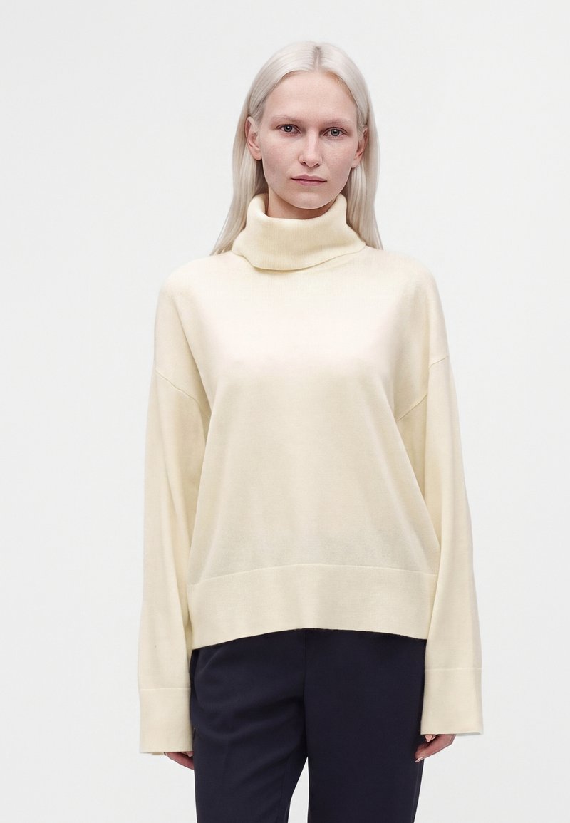 Femme à la peau pâle et aux cheveux blonds raides portant un pull à col roulé crème et un pantalon noir sur un fond clair uni.