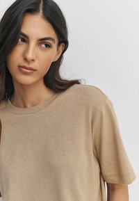 Beżowa koszulka z krótkim rękawem, z okrągłym dekoltem i miękkim, teksturowanym materiałem. Prosty fason o luźnym kroju, bez widocznych wzorów czy akcentów.