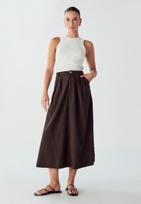 Brauer, plissierter, bodenlanger Rock mit Knopfschluss und Taschen, kombiniert mit einem weißen gerippten Tanktop und schwarzen flachen Sandalen.