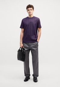 T-shirt viola a maniche corte con un motivo testurizzato, pantaloni grigi e una borsa di pelle nera. Il modello è in piedi contro uno sfondo neutro.