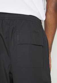 Närbild av en person som bär svarta shorts med resårmidja och en rektangulär fackficka på sidan, samt en vit skjorta instoppad.