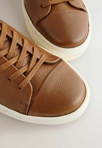 Zapatillas de cuero marrón con una superficie texturizada, puntera redondeada, cordones planos y suelas de goma blanca con detalles de diseño minimalistas.