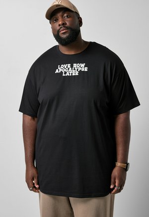 Uomo con barba che indossa un berretto beige, maglietta nera con testo bianco "LOVE NOW APOCALYPSE LATER,", pantaloni beige, orologio e anelli, in piedi davanti a uno sfondo grigio.