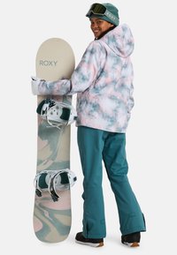 Roxy snowboard features a pastel marbled design with white, green, and pink. The rider wears a patterned jacket and teal pants, holding the board.