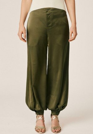 Mujer con pantalones sueltos verde oliva con puños fruncidos abotonados y sandalias doradas con tiras, de pie frente a un fondo liso.