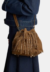 Borsa a secchiello in camoscio marrone con frange e borchie argentate, dotata di chiusura con coulisse e tracolla removibile.