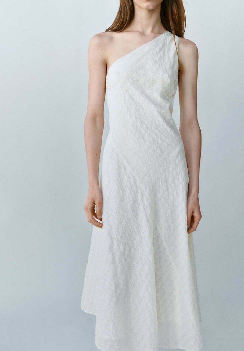 Femme portant une robe blanche texturée sans manches, à une épaule, debout devant un fond clair uni.