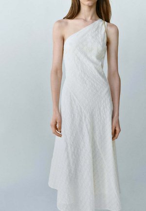 Femme portant une robe blanche texturée sans manches, à une épaule, debout devant un fond clair uni.