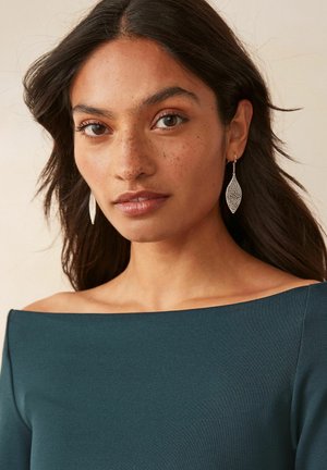Teal off-shoulder top with a smooth texture, highlighting a model's face adorned with silver leaf earrings. Subtle freckles visible.