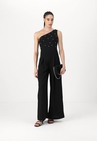 Black jumpsuit with one shoulder, embellished with white pearls, wide legs, and pleats. Holds a black clutch with a chain. Simple black heels.