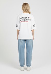 Bílý oversized tričko s černým a červeným textem na zádech, s grafikou srdcí na rukávech. V kombinaci s volnými, světle modrými džíny.