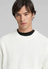 Maglione bianco lavorato a maglia con collo alto e rotondo. Presenta un motivo a rilievo e polsini a coste, indossato sopra una camicia nera.