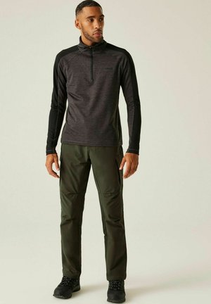 Homme debout portant un haut à manches longues noir et gris foncé avec fermeture éclair quart, un pantalon cargo vert olive et des chaussures de randonnée noires.