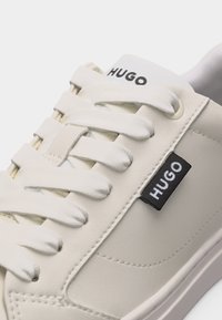 Balta sportinė avalynė su audinio raišteliais, ant liežuvėlio ir šoninės etiketės juodai užrašytas "HUGO" logotipas, lygi siūta viršutinė dalis, artimas vaizdas.