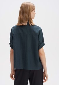 Maglietta corto maniche in verde scuro con una vestibilità rilassata. Presenta un tessuto texturizzato, maniche piegate e un dettaglio alla spalla posteriore.