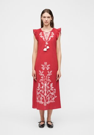 Moteris stovi apsirengusi raudona midi ilgio suknele su baltais gėlių siuvinėjimais, raukšlėtomis rankovėmis, kutelių raišteliais prie kaklo ir juodais Mary Jane stiliaus batais.