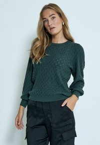 Maglione texture verde scuro con motivo a onde, polsini a coste e scollatura a girocollo, abbinato a pantaloni cargo neri.