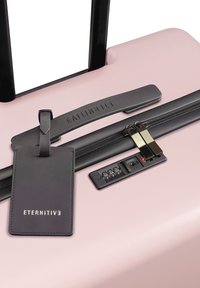 Jasnoróżowa walizka z czarnym teleskopowym uchwytem, zamkiem błyskawicznym i systemem zamka; zawiera czarny skórzany identyfikator bagażu z wytłoczonym napisem "ETERNITIV3".