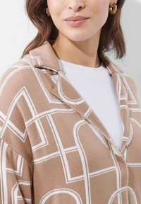 Tan gebreide cardigan met witte geometrische patronen. Voorzien van een ingesneden kraag en knopen, gedragen over een wit T-shirt. Textuurdetail van de stof zichtbaar.