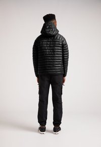 Giacca puffer nera con cappuccio, caratterizzata da un design trapuntato. Indossata con pantaloni cargo neri e scarpe sportive. Texture liscia, stile minimalista.