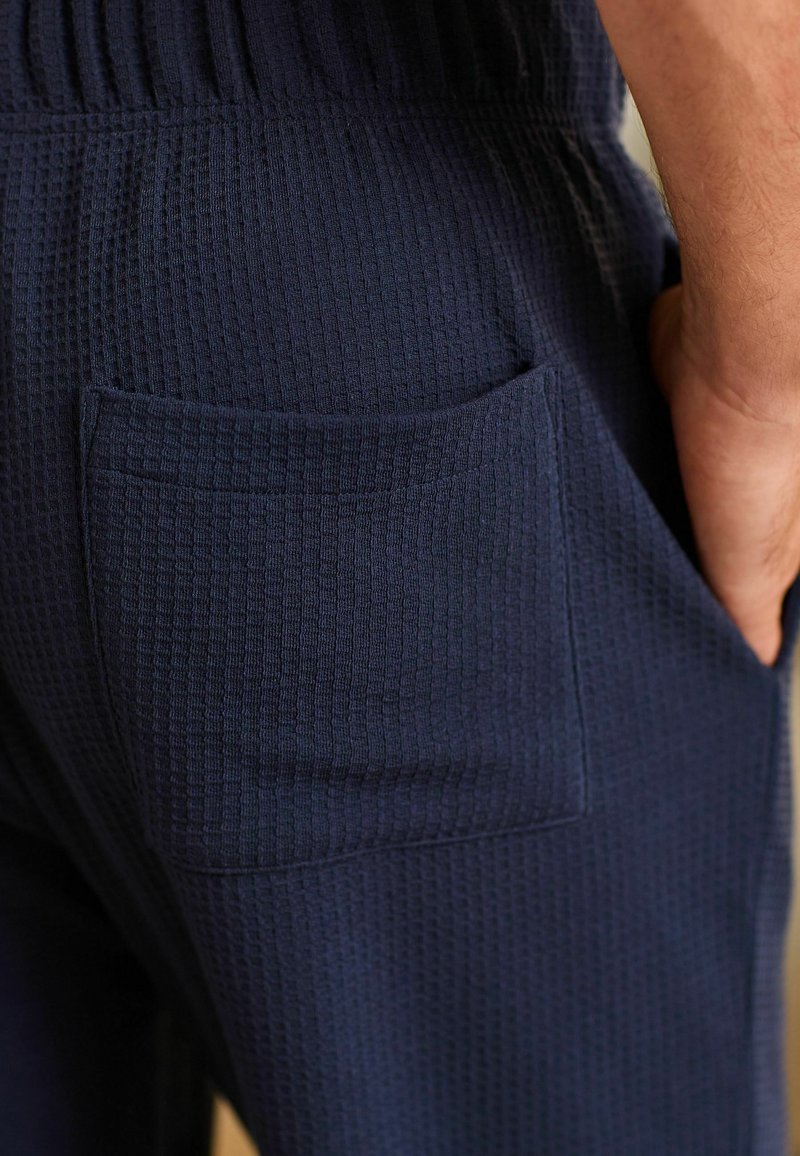 Dunkelblaue, strukturierte Hose mit sichtbarem Gesäßtaschen-Feature. Der Stoff hat ein waffelähnliches Muster und einen elastischen Bund für zusätzlichen Komfort.