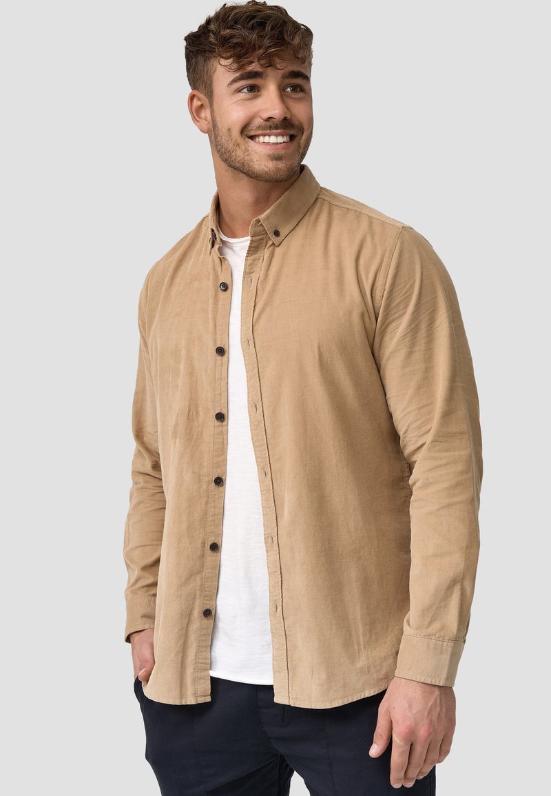 Camisa de pana marrón con botones, cuello de camisa y mangas largas, superpuesta sobre una camiseta blanca. Presenta un acabado texturizado y un corte relajado.