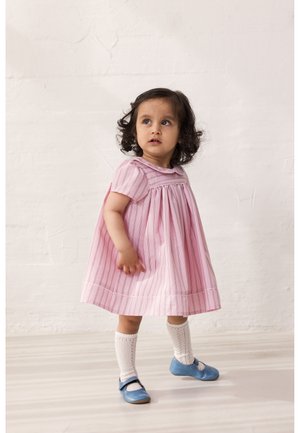 Νηπιακό κορίτσι με σγουρά σκούρα μαλλιά, φορώντας ροζ ριγέ φόρεμα, λευκές κάλτσες έως το γόνατο και μπλε παπούτσια Mary Jane, στέκεται μπροστά σε έναν απλό τοίχο.