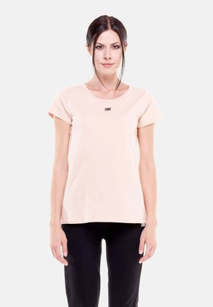 Jeune femme aux cheveux foncés attachés, portant une chemise à manches courtes beige clair et un pantalon noir, debout devant un fond uni.