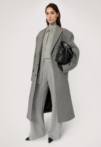 Szary wełniany płaszcz, zestaw szarych swetra i spodni, pasek w paski, czarna skórzana torba, czarne szpiczaste buty. Teksturowana tkanina i dopasowany krój.