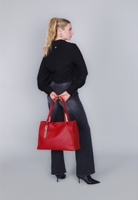 Rode leren tote bag met dubbele handvaten, voorzien van een metallic "CAFÉ NOIR" accent, weergegeven tegen een grijze achtergrond met een model.