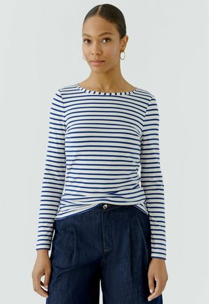 Frau mit zurückgebundenem Haar, die goldene Creolen trägt, ein weiß-blau gestreiftes langärmeliges Hemd und dunkelblaue Hose vor einfarbigem Hintergrund.