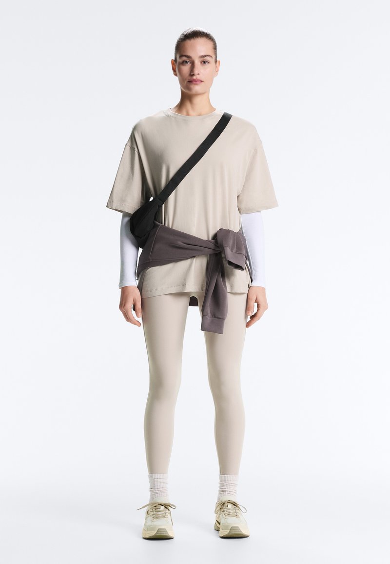 Frau, die ein beige oversized T-Shirt, beige Leggings, weißes langärmeliges Unterhemd, beige Sneaker, einen dunklen Sweatshirt, der an der Taille gebunden ist, und eine schwarze Umhängetasche trägt.
