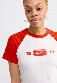 Balts īsām piedurkņu T-krekls ar sarkaniem raglan piedurkņu un retro Nike logo, kas izdrukāts sarkanā krāsā, zem logo atrodas skaitļi "19 72".
