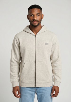 Homme portant un sweat à capuche zippé beige clair avec poches avant et un jean bleu clair, debout devant un fond gris uni.