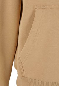 Materialul de hoodie bej are o textură vizibilă, având un buzunar tip kangur și o talie cu guler ribbed. Detaliile de cusătură sunt, de asemenea, proeminente.