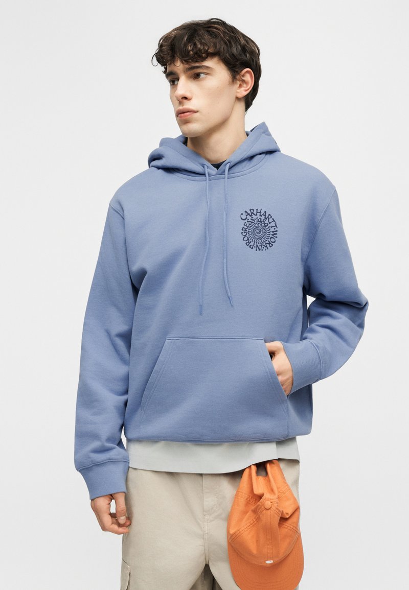 Joven con sudadera azul Carhartt con bolsillo frontal, sosteniendo una gorra naranja en el bolsillo de sus pantalones beige, mirando hacia la izquierda sobre un fondo blanco.