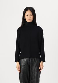 Svart turtleneck genser i ribbestrikk, med en avslappet passform og sidesplitt, sammen med skinnende svarte plissert bukser.