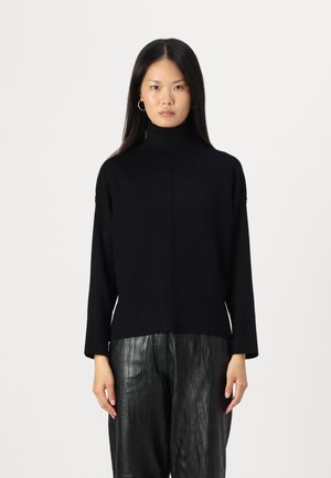 Pull à col roulé noir en tricot côtelé, avec une coupe décontractée et des fentes sur les côtés, associé à un pantalon plissé noir brillant.