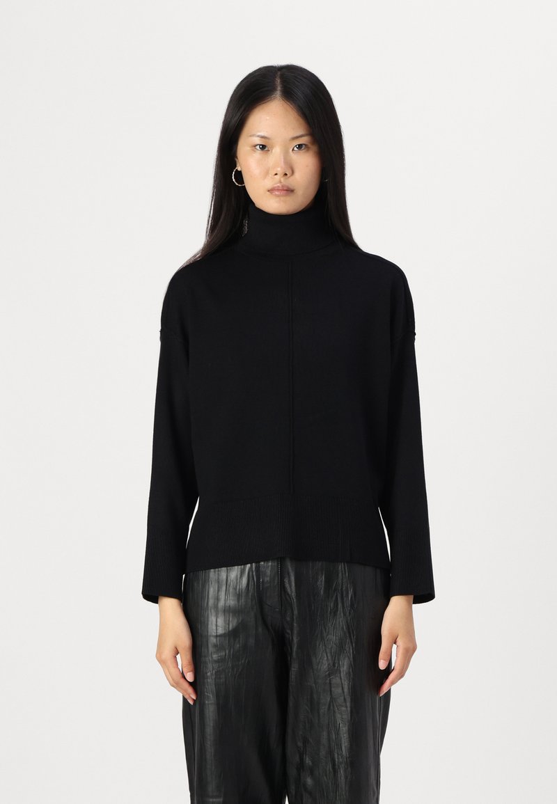 Svart turtleneck genser i ribbestrikk, med en avslappet passform og sidesplitt, sammen med skinnende svarte plissert bukser.