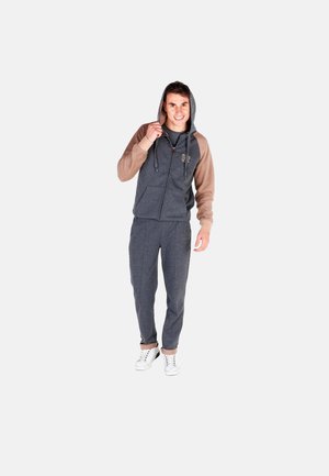 Jeune homme en survêtement gris avec des manches marron et des baskets blanches, souriant et tenant la capuche de sa veste.